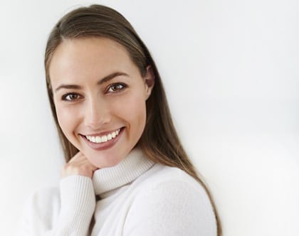 Head-and-shoulders photo of a young brunette woman with long hair wearing a white turtleneck sweater smiling and with her right hand under her face, for examples of beautiful smile makeovers with porcelain veneers from Kentucky Dental Group of Lexington.